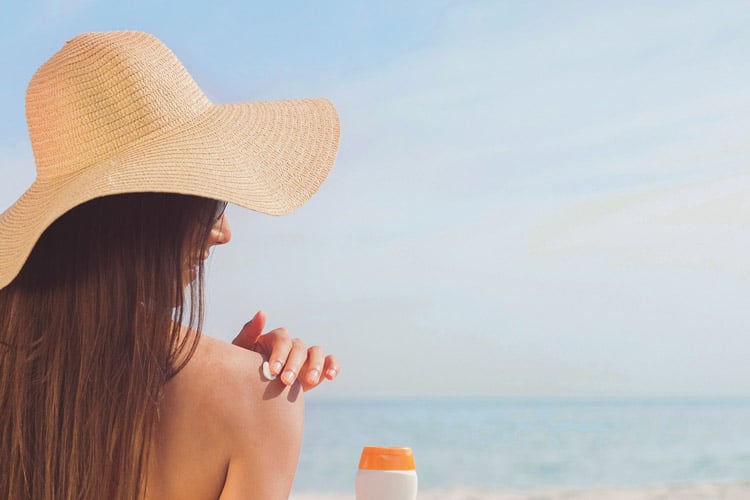 Frau am Strand mit Sonnenhut cremt sich mit Sonnencreme ein